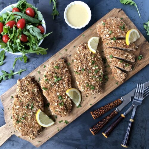 Salad, dip, lemons and almond crusted chicken served on a wood board.