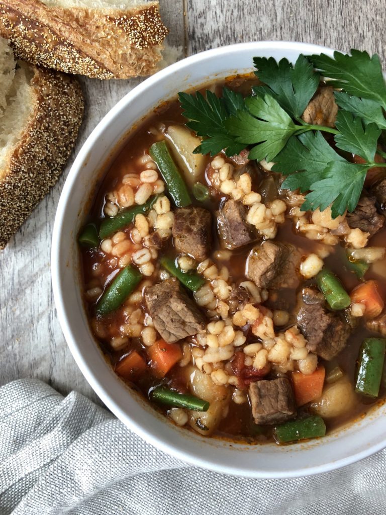 Top down view beef barley soup bowl with Italian bread on side.