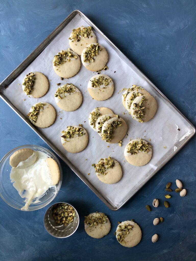 Vanilla cookies being dipped in chocolate with pistachio sprinkle.
