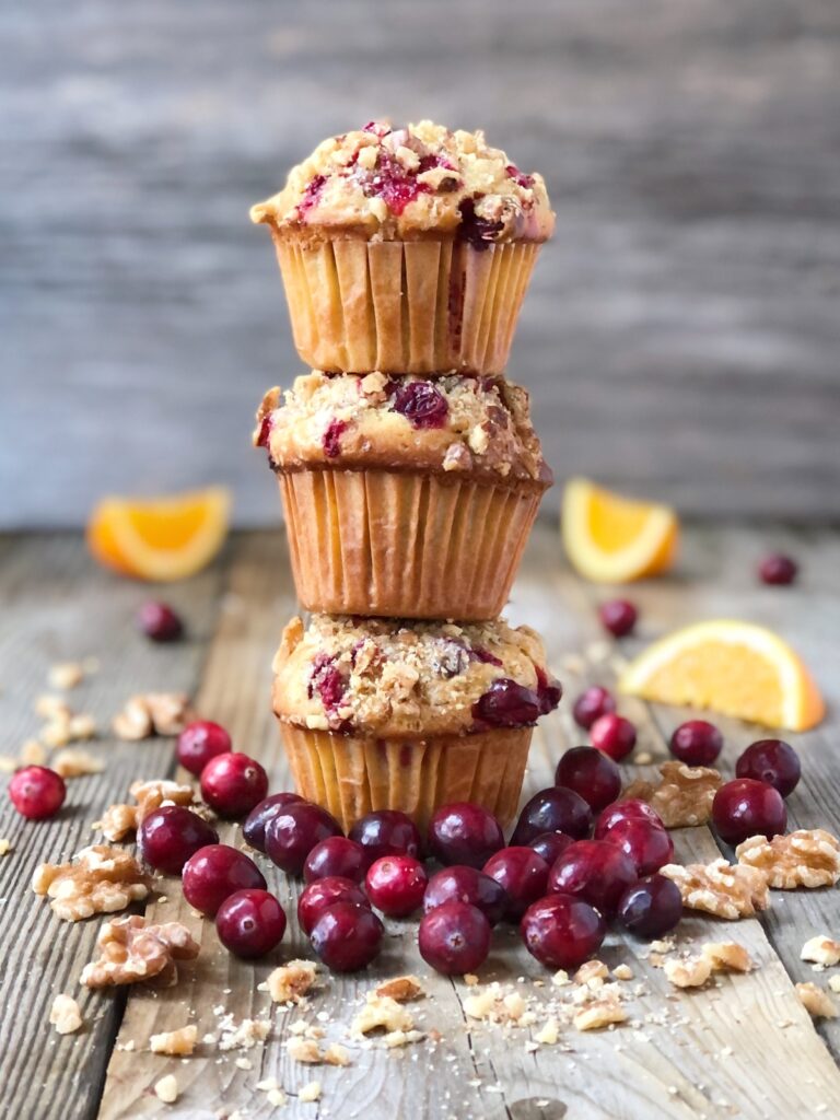 Three cranberry muffins stacked on a wood board with cranberries, walnuts and orange slices around them.