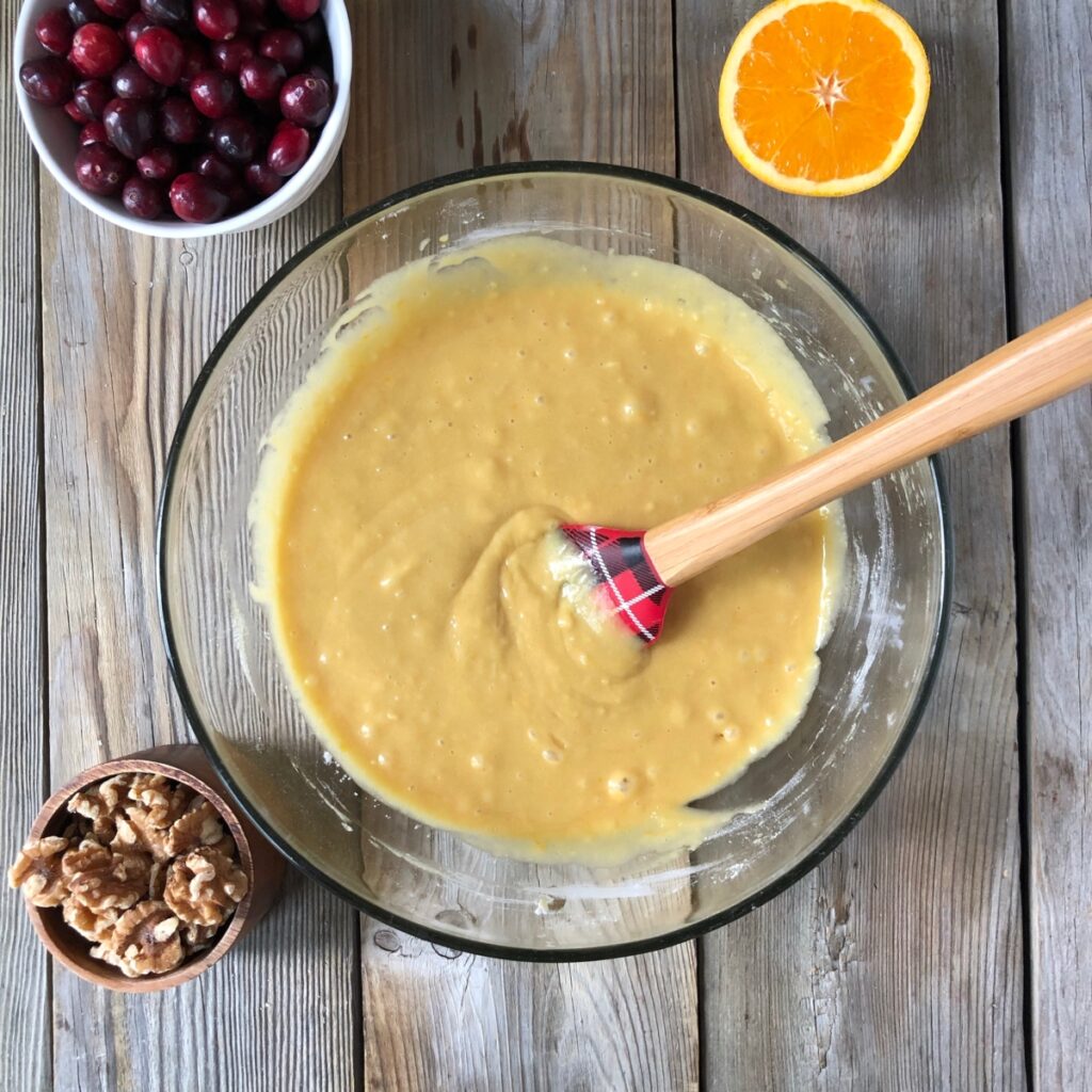 Muffin batter mixed in clear bowl with spatula in it and orange, cranberry, walnuts around it.
