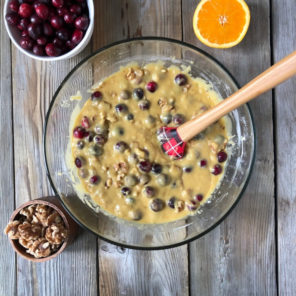 Muffin batter with cranberries and walnuts added with spatula stirring, orange, cranberry walnuts beside it.