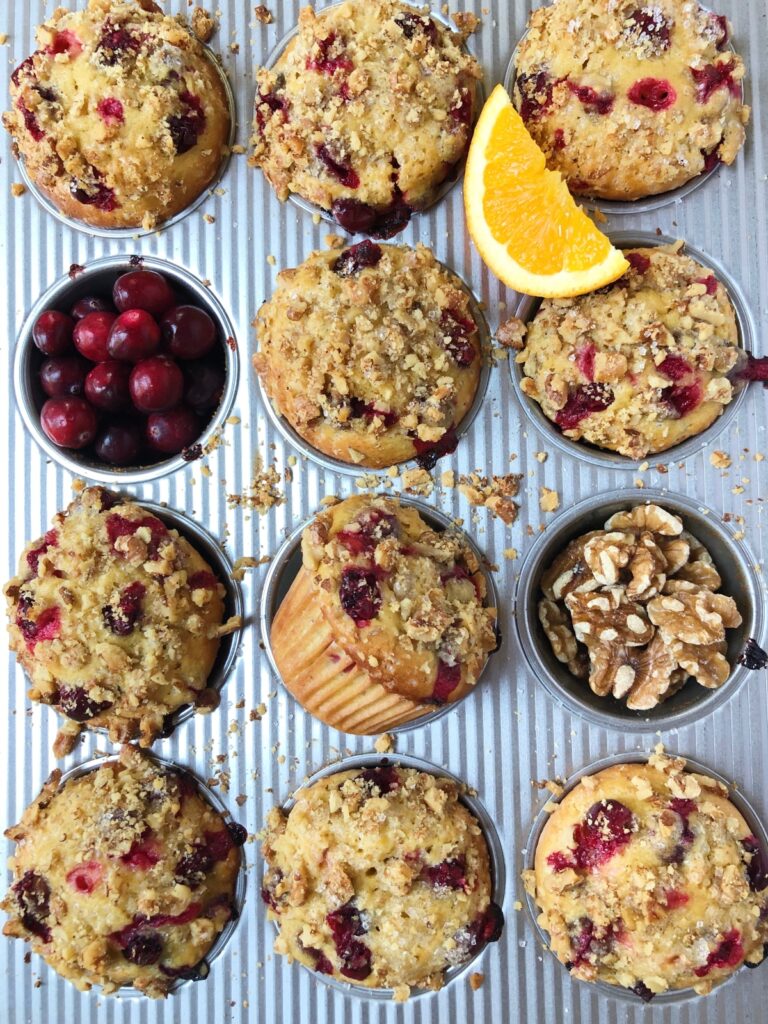 Top down close view of cranberry orange muffins in muffin tin with oranges, cranberries and walnut crumbs on tin.