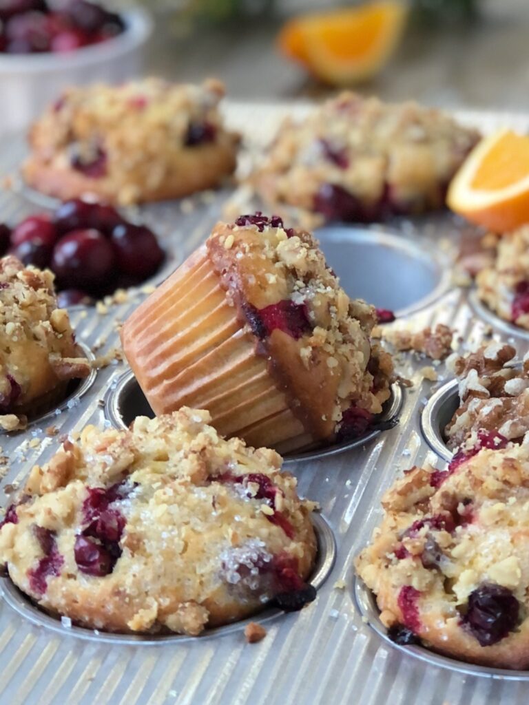 Close view, few muffins in muffin tin with one popping up on angle, oranges, cranberries, walnuts scattered.