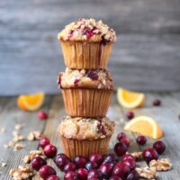 Three cranberry orange muffins stacked on wood board with cranberries, walnuts and orange slices scattered