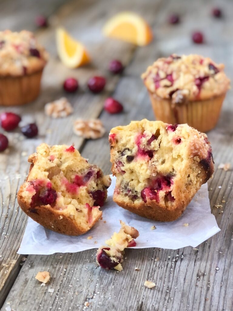 One cranberry orange muffin split open on parchment, 2 muffins behind with cranberries, walnuts, orange slices scattered.