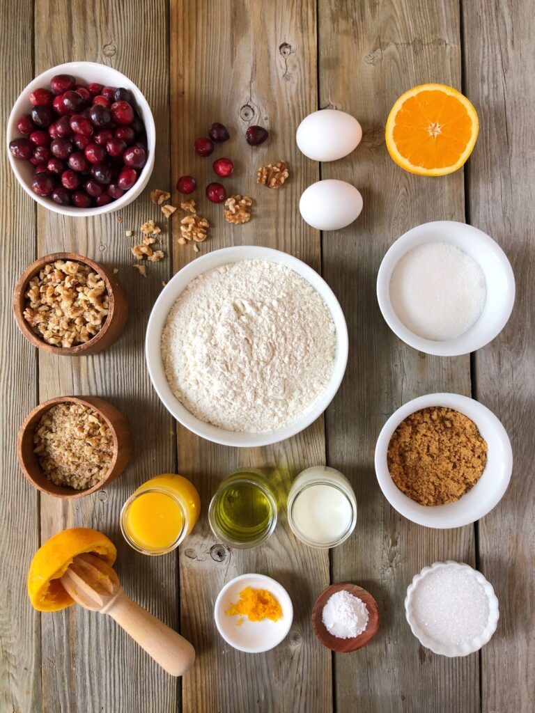 Essential ingredients eggs, oil, milk, oranges, sugar, flour, cranberries, walnuts in bowls laid out on wood board.