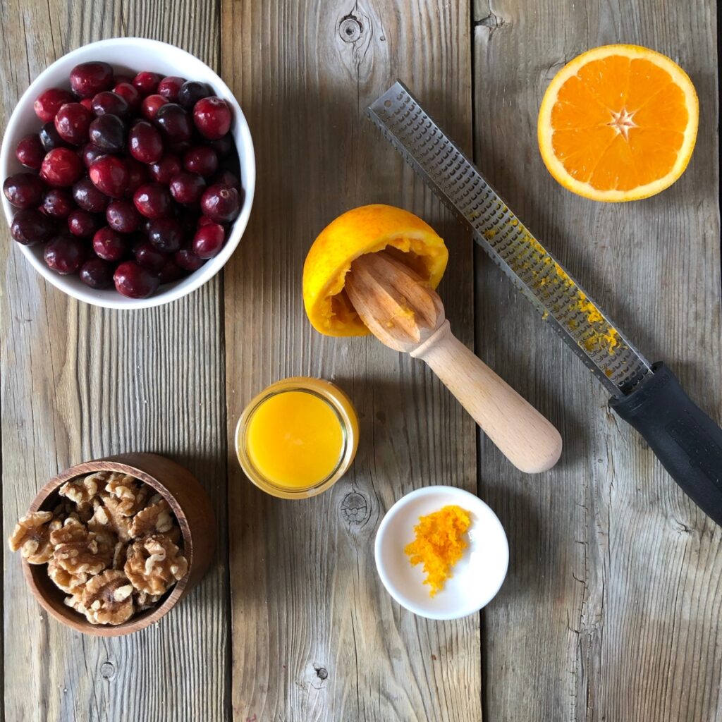Zested orange with rasp and juiced orange, wood juicer on wood board, orange cranberry walnuts around it.