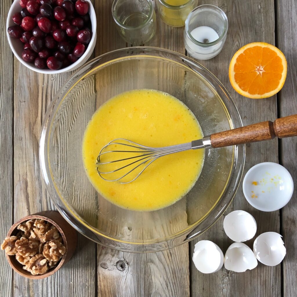 Clear bowl with whisk whisking wet ingredients surrounded by cracked eggs, orange, cranberry, walnuts.