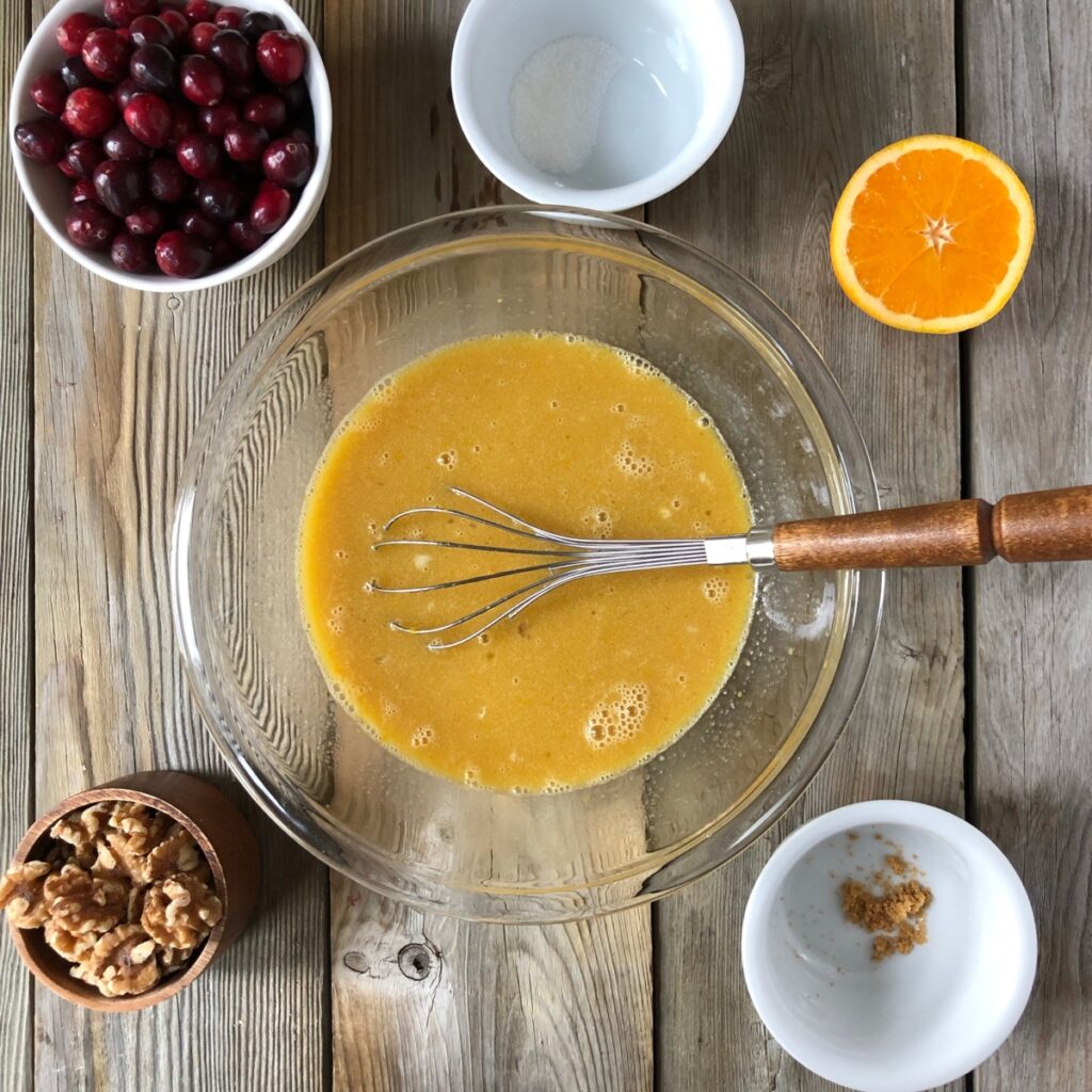 Clear bowl with whisk whisking sugars to wet ingredients surrounded by orange, cranberry, walnuts.