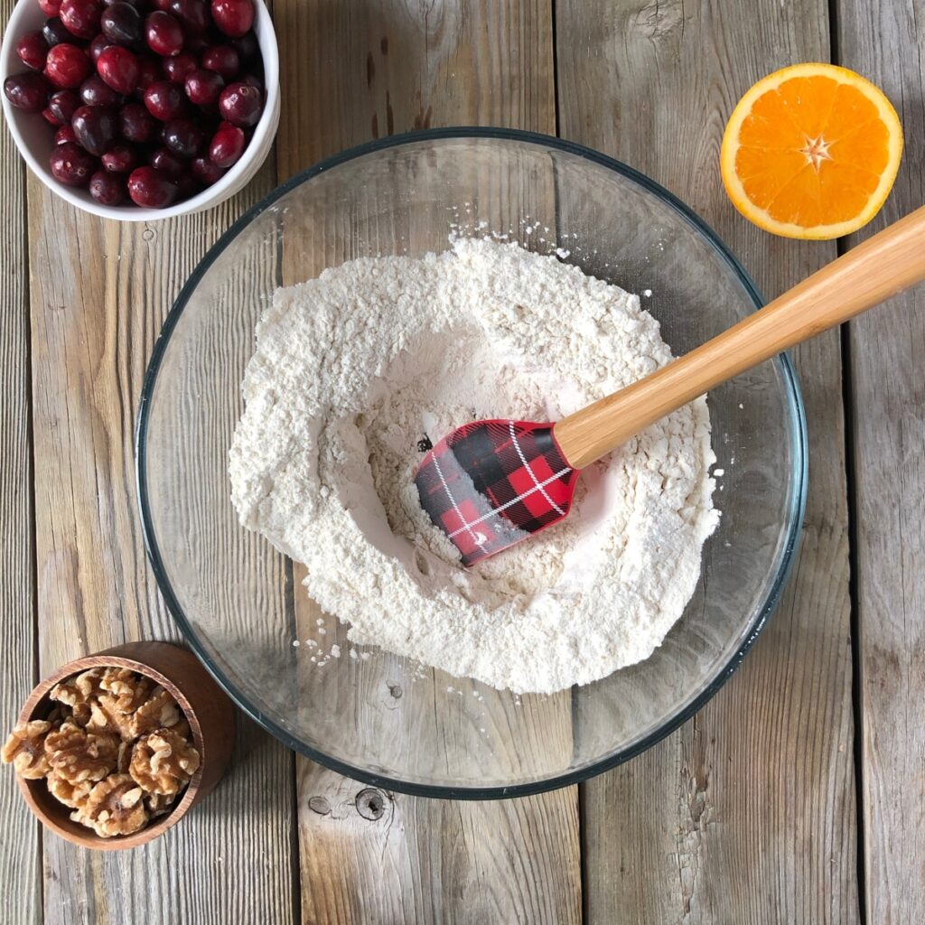 Clear bowl with spatula stirring dry flour ingredients with orange, cranberry, walnuts beside it.