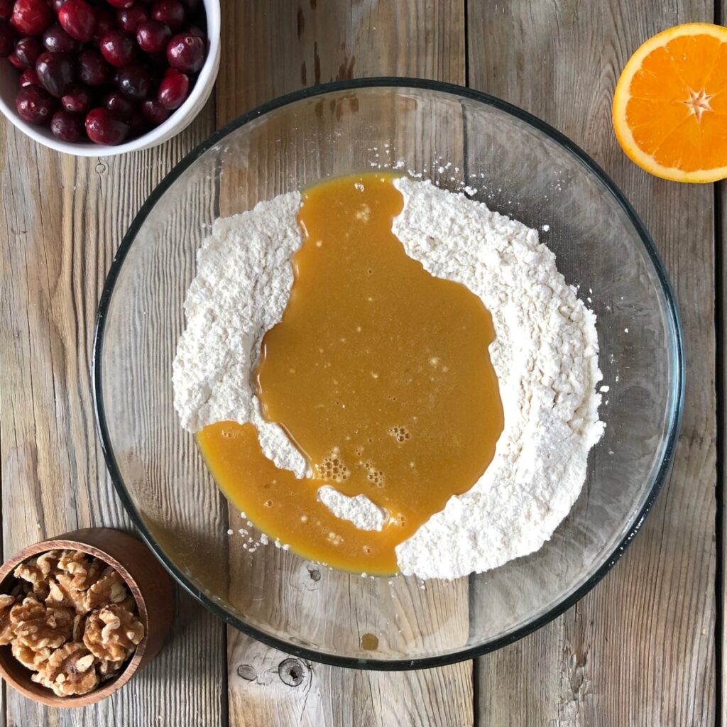 Adding wet ingredients to clear bowl with dry ingredients, spatula in bowl, orange, cranberry, walnuts near it.