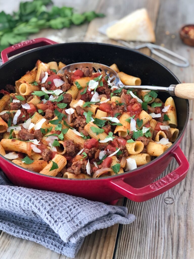 Sausage pasta in skillet on tea towel with serving spoon and parmesan cheese.