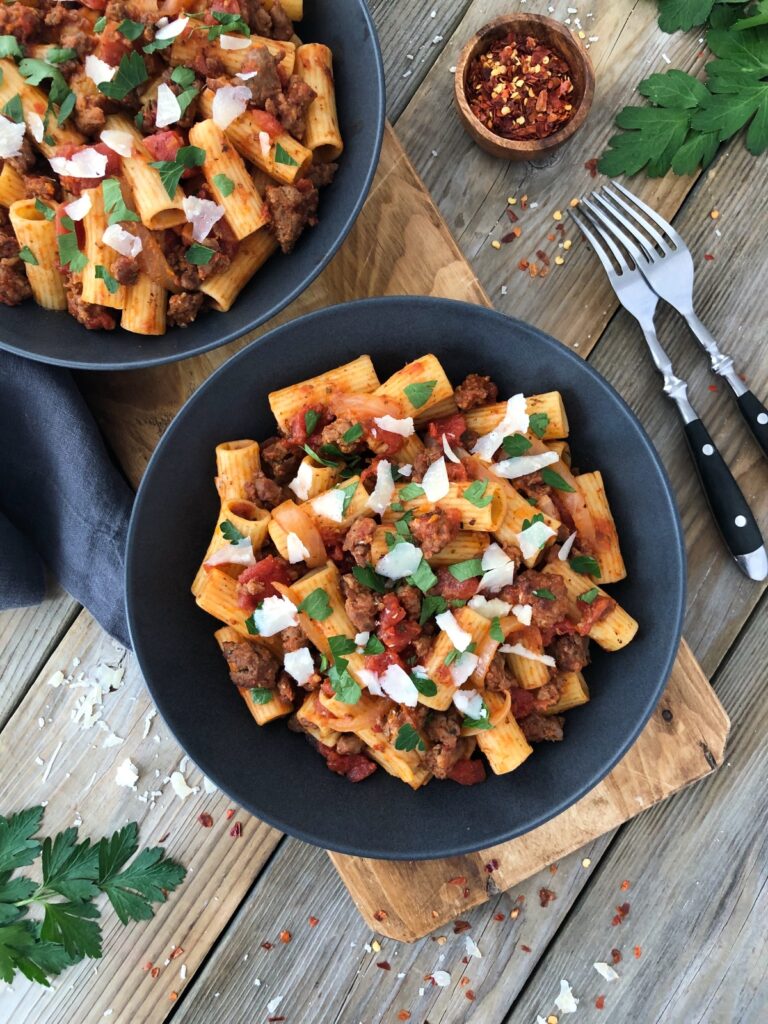 Top down view two full bowls of italian sausage pasta with two forks, chili and parsley garnish