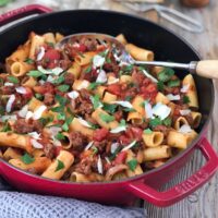 Sausage pasta in skillet on grey tea towel garnished with parmesan and parsley with serving spoon