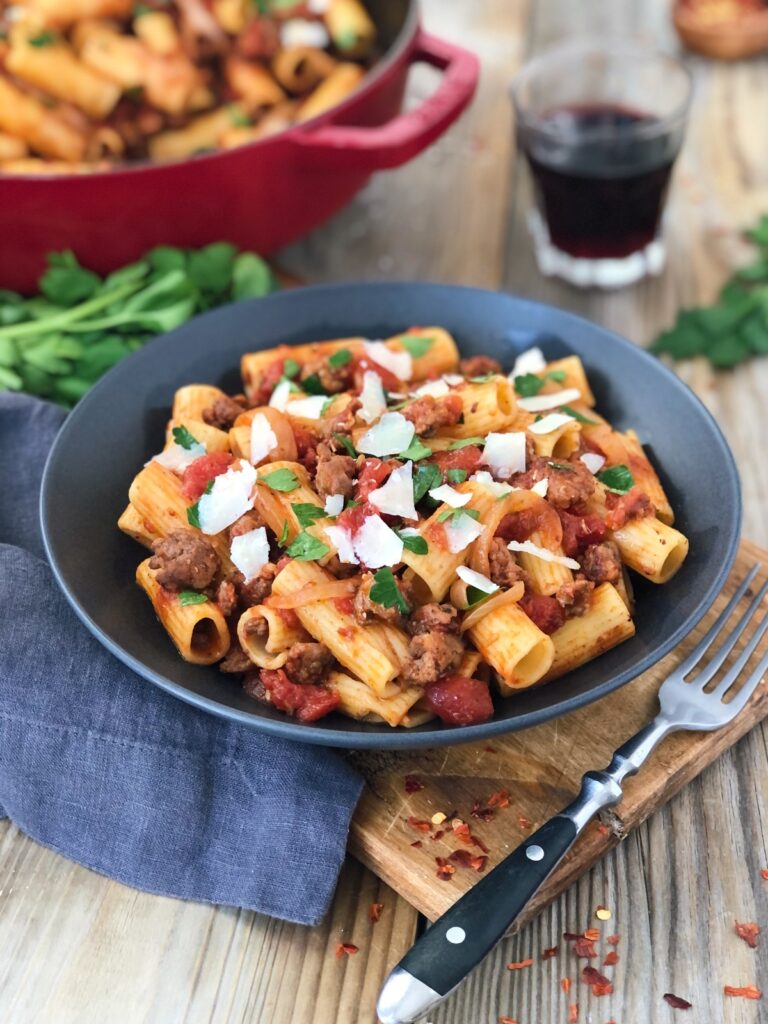 Pasta serving in bowl on a board with cloth napkin, fork on side with glass of red wine.