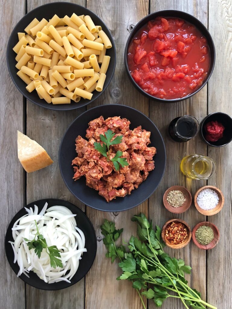 Pasta, tomatoes, parmesan cheese, wine, olive oil, onions, seasonings and parsley laid out on a board.