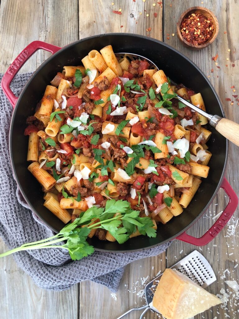 Skillet with sausage rigatoni with serving spoon garnished with parmesan, parsley and chili