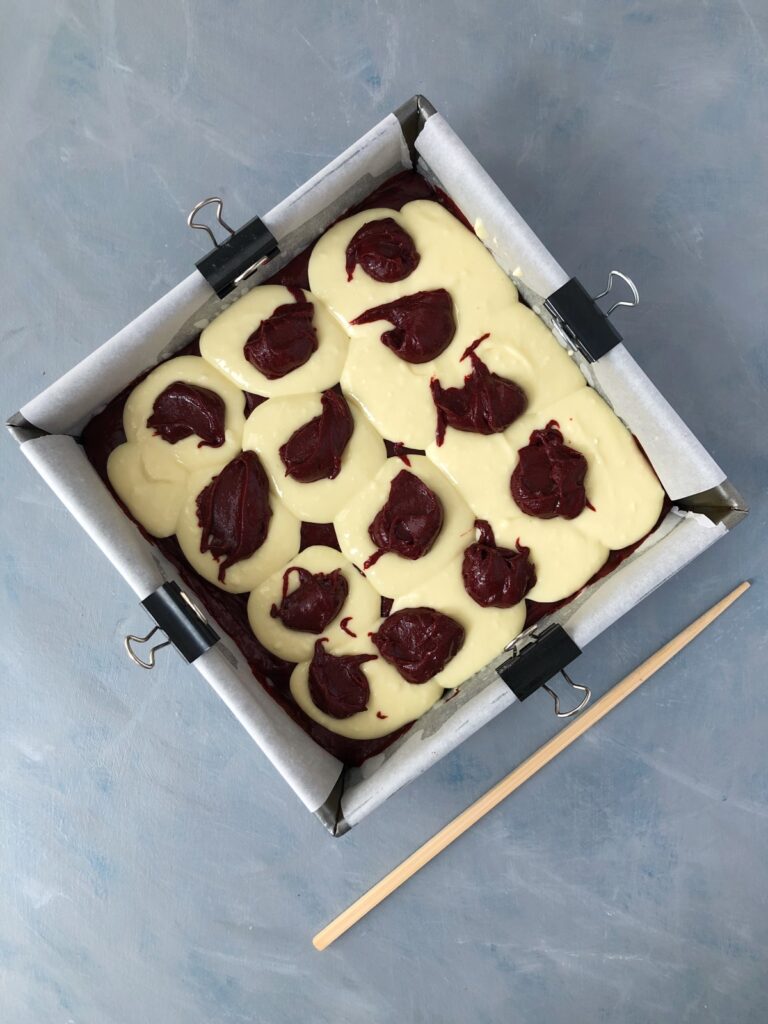 Cheesecake batter dolloped over brownie batter in baking pan.