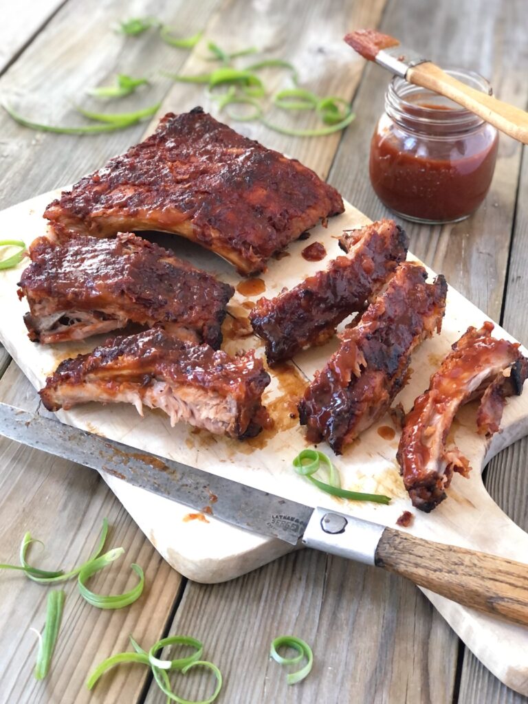 Tender cooked ribs enrobed in sauce being sliced with a knife on a wood board.