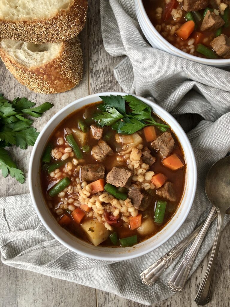 Two bowls beef barley soup servings with spoons and bread.