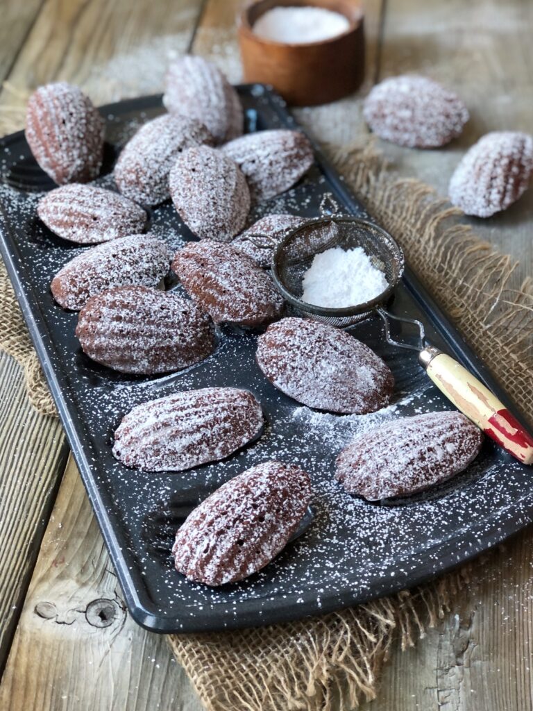 Madeleines on baking pan with a sieve being dusted with icing sugar.