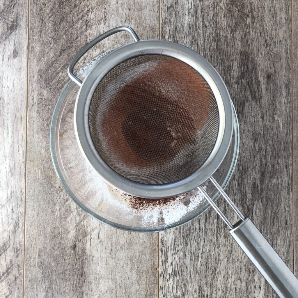 Flour and cocoa powder being sifted into a bowl with a sieve.