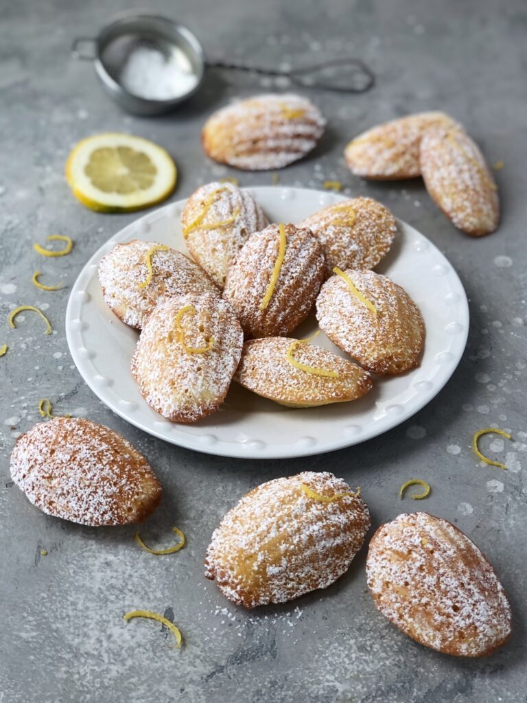 Lemon madeleines on a plate and some scattered on table.