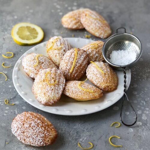 Lemon madeleines on a white plate dusted with icing sugar.