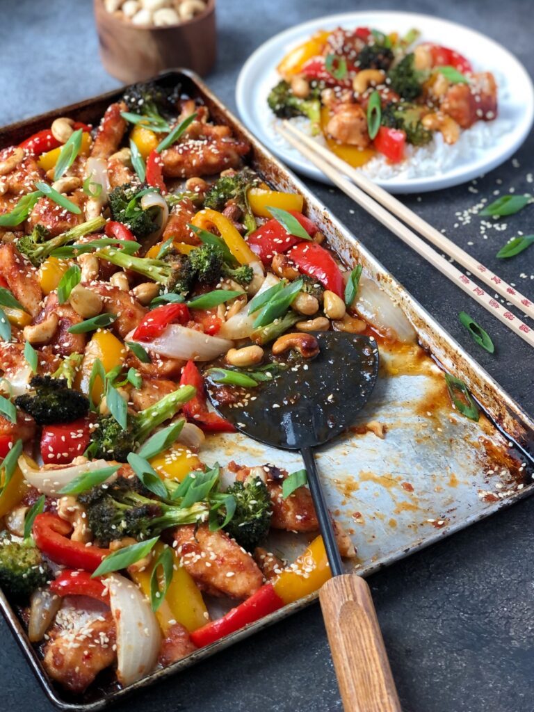 Flat scoop dishing out portion of cashew chicken and vegetables from sheet pan.