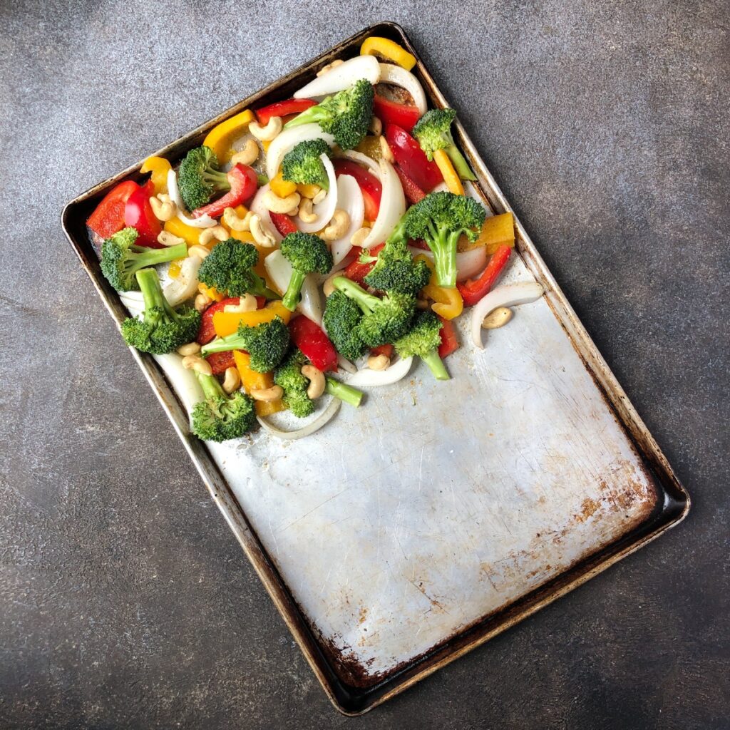 Broccoli, onion, peppers and cashews arranged on half of a sheet pan.