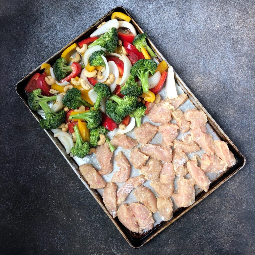 Coated chicken arranged on pan beside vegetables and cashews.